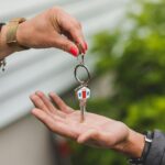 Novations vs. Wholesaling: Which Real Estate Strategy Is For You? Close-up of a hand handing over a key with a house keychain, symbolizing real estate transaction.